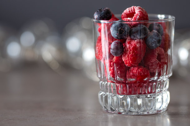 桌上放着一只玻璃杯，里面装满了冰冻的覆盆子和蓝莓。图片下载