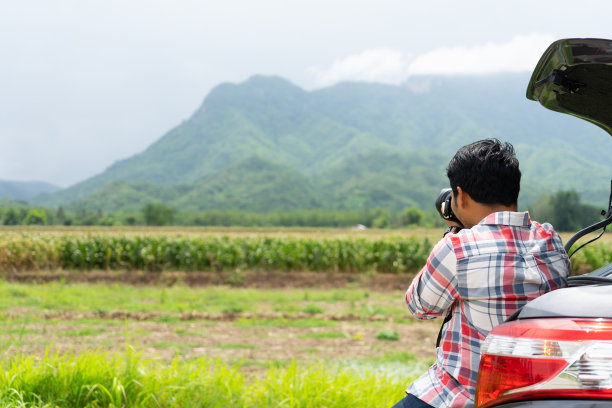 一名年轻人坐在汽车后备箱里，用单反相机以绿色的玉米地和山区自然为背景拍照。暑假与汽车上的道路概念。图片下载