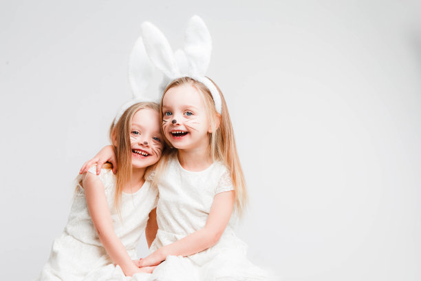 一对金发的双胞胎，穿着白裙子，长着兔耳朵。灰色背景上的工作室照片。孩子们庆祝复活节。图片下载