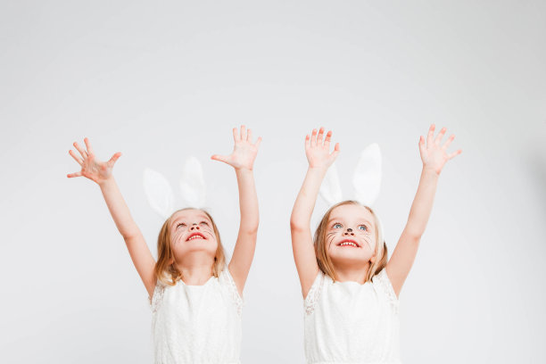一对金发的双胞胎，穿着白裙子，长着兔耳朵。灰色背景上的工作室照片。孩子们庆祝复活节。图片下载
