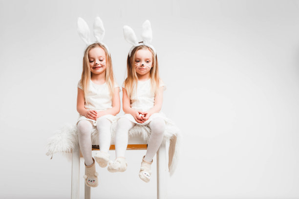 一对金发的双胞胎，穿着白裙子，长着兔耳朵。灰色背景上的工作室照片。孩子们庆祝复活节。图片下载