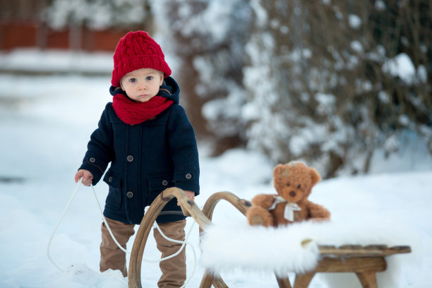 宝宝在雪地里玩泰迪熊，冬天的时候。穿着蓝色外套的蹒跚学步的小男孩，在雪橇上滑行，抱着泰迪熊，在冬季公园的户外玩耍图片下载