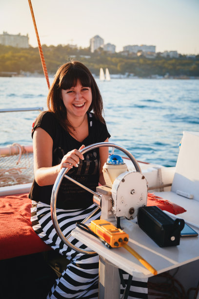年轻漂亮的微笑女人在黑色衬衫和条纹裙子驾驶豪华游艇在海上，日落图片下载