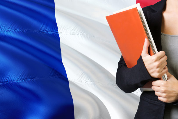 学习法语语言概念。一名年轻女子站在法国国旗后面。老师拿着书，橘黄色的空白书封面。图片下载