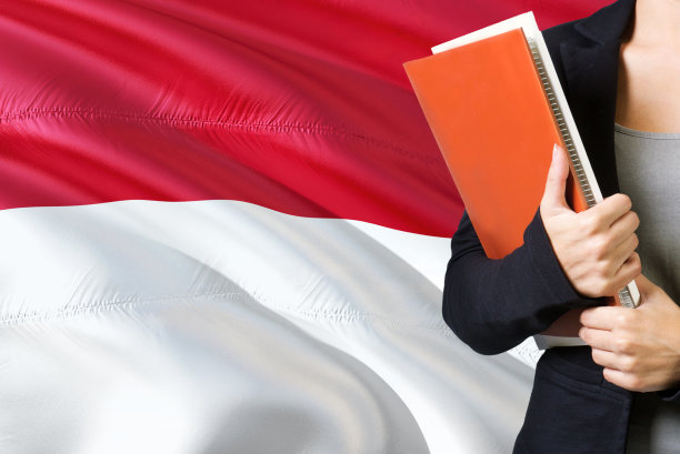 学习印尼语概念。一名年轻女子站在印度尼西亚国旗的背景下。老师拿着书，橘黄色的空白书封面。图片下载