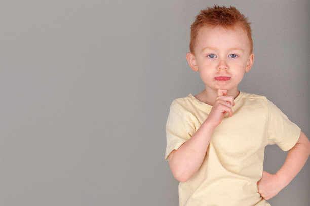 火红头发的年轻男孩在黄色衬衫在手指到下巴在深刻的思考姿态在风景格式在灰色背景与复制空间。隐喻思维。表情包和可爱红头发的海报图片。图片下载