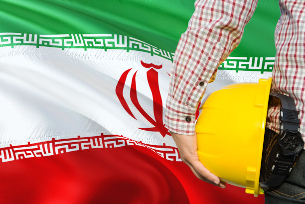 伊朗工程师手持黄色安全帽，背景是挥舞的伊朗国旗。建筑施工概念。图片下载