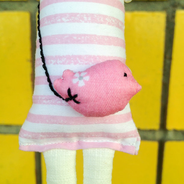 一个手工娃娃的特写镜头，穿着一件粉红色和白色的条纹连衣裙，用线把一只粉红色的鸟攥在手里，靠在一堵黄色瓷砖墙上图片下载