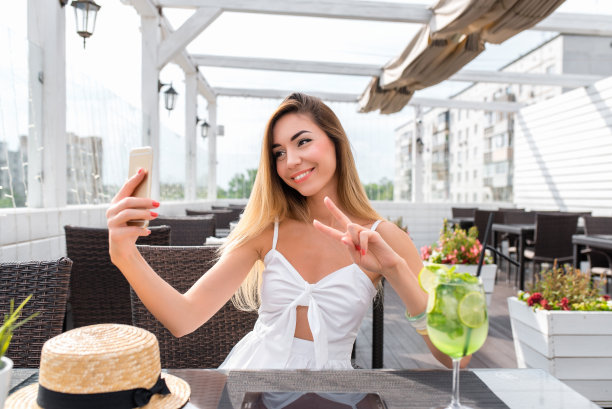 一个漂亮的女孩坐在阳台上的咖啡馆里。在夏天的一个阳光明媚的日子里。照片在手机上，幸福的微笑。在智能手机的视频通话中，摄像头上的姿势传输被保存下来。图片下载