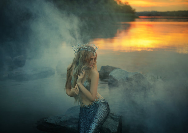 美丽的海公主谦虚地向一边看，梳着她金色的长发，可爱的贝壳头饰配上银色的尾巴，迷人的仙女在河岸边，阳光在浓雾中突出图片下载