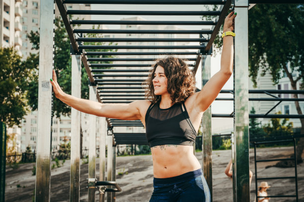 主题是妇女运动与健康。美丽的白种女人与卷长发摆姿势在户外运动场地抱着单杠，腹部纹身在运动上衣和紧身裤图片下载