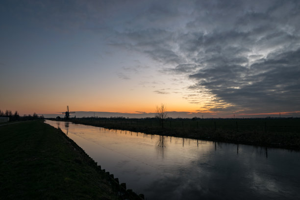 一条荷兰运河被冰覆盖着。傍晚天空衬托出远处风车的轮廓。图片下载
