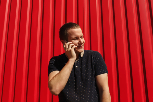 英俊的成年男子使用他的手机，微笑在红色的背景和自由的空间。图片的快乐的年轻人站在红色的墙背景与手机图片下载