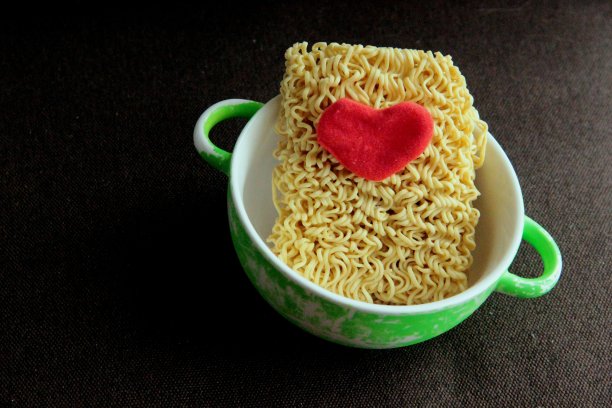 绿色破旧的盖碗，即食的黄色干面和红色的心绒织物在棕色的背景上近距离模糊。图片下载