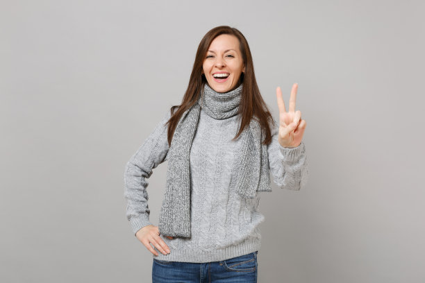 快乐的年轻女子在灰色毛衣，围巾显示胜利标志孤立在工作室的灰色墙壁背景。健康时尚的生活方式，人们真诚的情感，寒冷的季节理念。模拟拷贝空间。图片下载