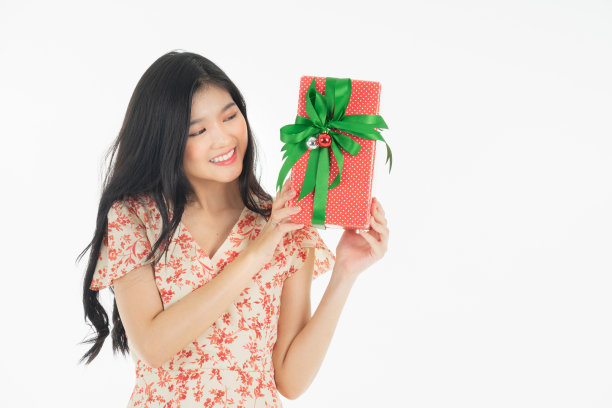 亚洲好奇的女人在红色裙子庆祝她的生日或新年礼盒的照片。年轻女子拿着礼盒与红色的蝴蝶结正在兴奋和惊讶的节日现在孤立的白色背景图片下载