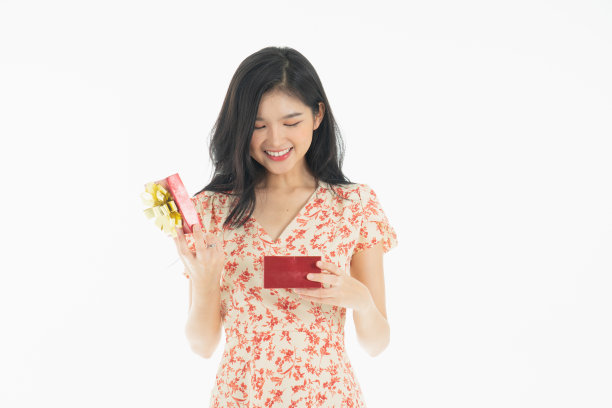 亚洲好奇的女人在红色裙子庆祝她的生日或新年礼盒的照片。年轻女子拿着礼盒与红色的蝴蝶结正在兴奋和惊讶的节日现在孤立的白色背景图片下载