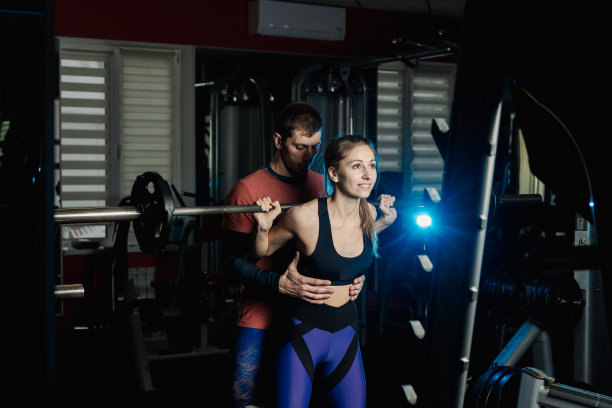 在体育馆里，英俊的运动员用肩膀上的杠铃帮助一个漂亮的女孩下蹲图片下载