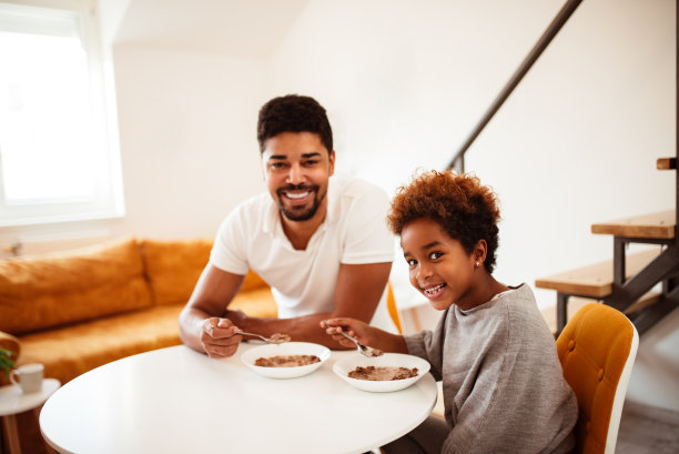 一个微笑的混血父亲和女儿的肖像吃谷类早餐，看着镜头。图片下载