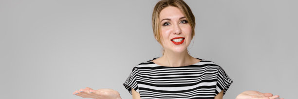 迷人的年轻金发女孩在条纹衬衫微笑与张开的双手站在灰色的背景图片下载
