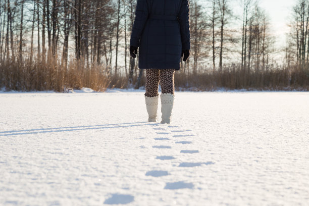 年轻的成年女子穿着白色的靴子在冰封的湖面上行走。人类身后的新脚印。享受在阳光明媚，寒冷冬日的森林漫步。后视图。图片下载