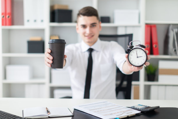 一个年轻人坐在办公室的一张桌子旁，手里拿着一杯咖啡和一个闹钟。照片景深，突出焦点玻璃和闹钟。图片下载