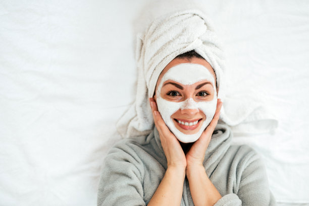 美丽的年轻女子躺在床上，脸上涂着乳脂或面膜。俯视图，看摄像头。图片下载