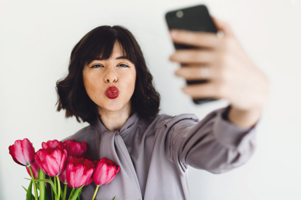 美丽快乐的女孩与郁金香花束和礼盒在室内白色背景，文字空间自拍。带着鲜花和手机的时髦年轻女子。国际妇女日图片下载