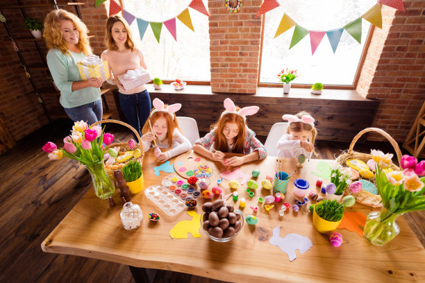 近距离的照片三个孩子制作装饰鸡蛋复活节标志孩子日两个妈妈带来礼盒看起来高兴可爱甜蜜的样子桌子满工艺坐大木头桌子地板图片下载