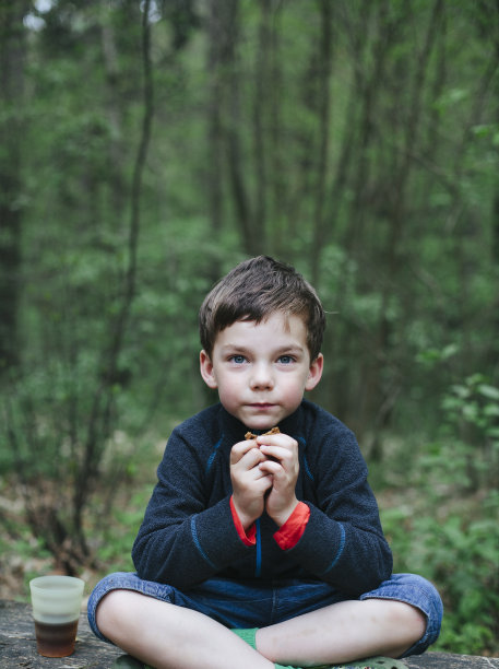 小男孩盘腿坐在长椅上，看着镜头，吃着他的面包在森林里的野餐午餐。图片下载