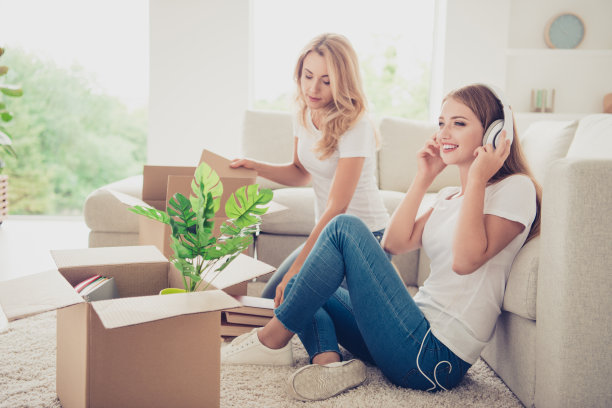 特写照片两个人妈妈和十几岁的女儿不关心帮助打开箱子听音频耳罩，而是穿着白色t恤牛仔裤在明亮的平坐在舒适的沙发地板上图片下载