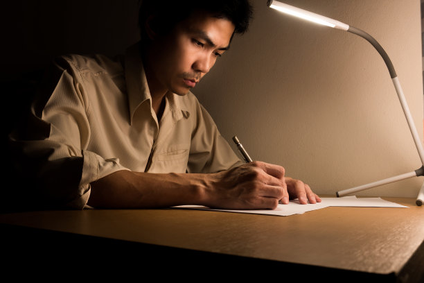 一个忙碌的商人晚上在办公室加班，正坐在椅子上。他使用一个led灯照明他的书桌，为写作和打字笔记本电脑研究信息图片下载