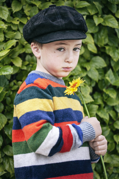悲伤，可爱的小男孩在彩虹五彩条纹毛衣和黑色绒面革报童帽拿着一个橙色的雏菊花图片下载