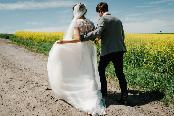 新娘、新郎走回春日的油菜花田、油菜花田。一对新婚夫妇在乡间直路上跑步去度蜜月。后视图。图片下载