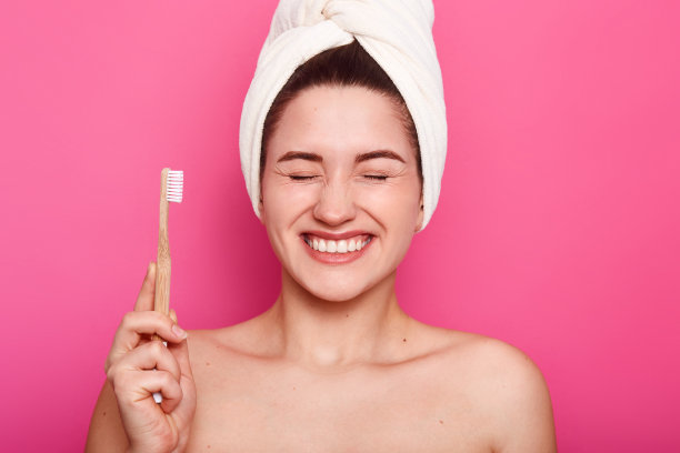 正面女性的特写镜头有光滑的皮肤，保持眼睛闭着，显示白色的牙齿，摆出的姿势，有白色的毛巾在头上，手持牙刷，准备刷牙，孤立的粉红色图片下载
