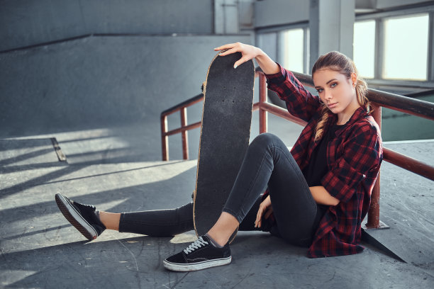 一个漂亮的年轻女孩穿着格子衬衫拿着一个滑板，而坐在旁边的研磨栏杆在滑板公园室内。图片下载