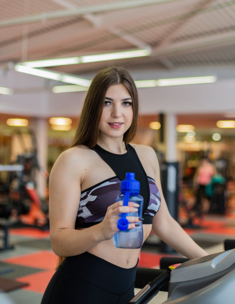 健身房的女人在锻炼喝水，开心地站在月球漫步者健身机器旁。美丽适合年轻的混血白种人亚裔女性健身模特在健身中心内图片下载