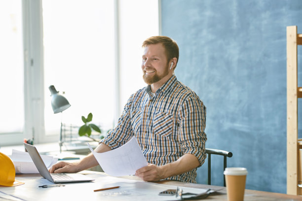 乐观兴奋英俊的年轻工程师戴着无线耳机坐在自己的工作室的桌子上，使用笔记本电脑，同时检查蓝图图片下载