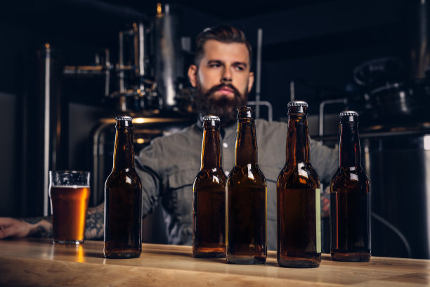 照片中前景是手工啤酒瓶，背景是正在喝酒的大胡子男子。图片下载