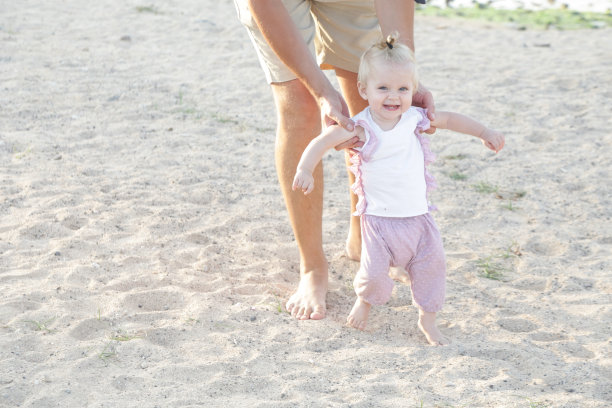 父亲帮助他的宝宝第一步，教宝宝走路的概念，户外偷拍照片在海滩上，花一天在海滩上，健康的家庭生活方式图片下载