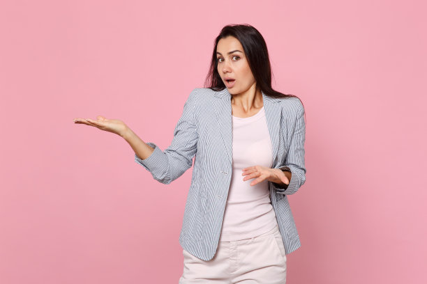 震惊的年轻女子在条纹夹克保持大张的嘴，指着分开的手在粉色粉彩墙背景在工作室。人真诚情感，生活理念。模拟拷贝空间。图片下载