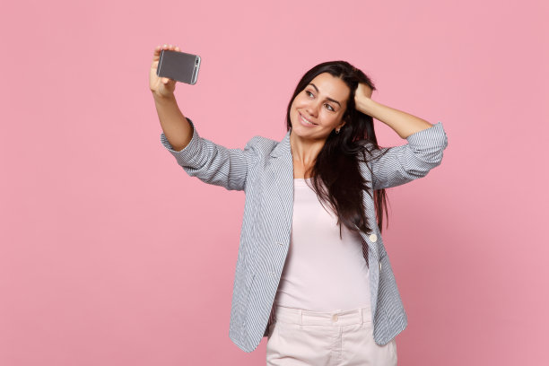 迷人的年轻女子在条纹夹克把手放在头上，在手机上做自拍孤立在粉色粉笔墙背景。人真诚情感，生活理念。模拟拷贝空间。图片下载