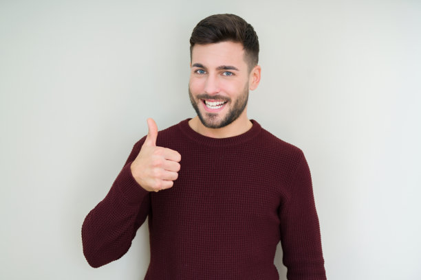 年轻英俊的男子穿着毛衣在孤立的背景做快乐的拇指手势与手。赞许的表情看着镜头，表示成功。图片下载