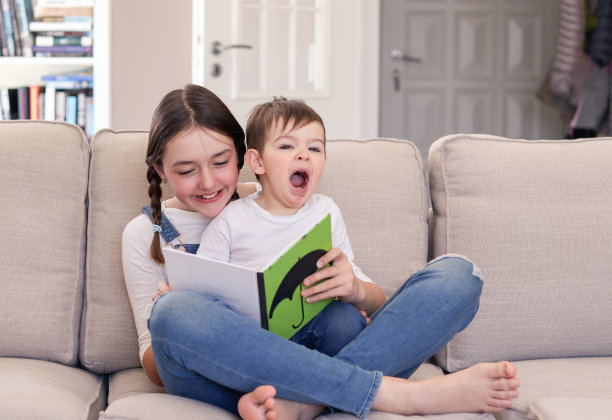 微笑的少女读书给她可爱的小弟弟坐在她的腿上，打哈欠在沙发上在家。兄弟姐妹关心和爱。姐妹和兄弟的优质时间。累了睡着的孩子图片下载