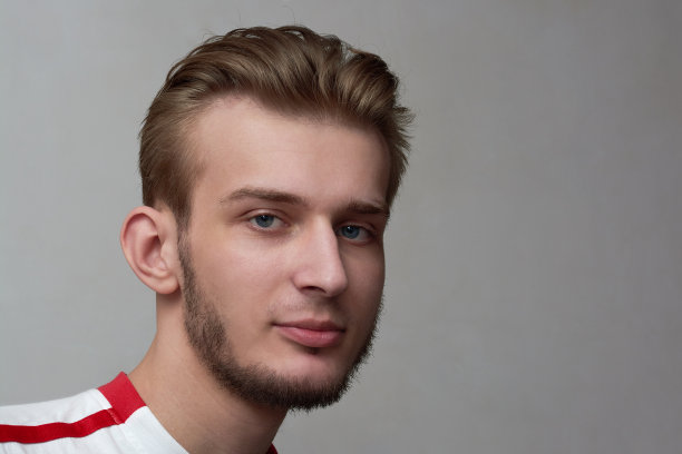 年轻男子的肖像带着淡淡的微笑。灰墙背景上留着胡子和时髦发型的男孩。图片下载