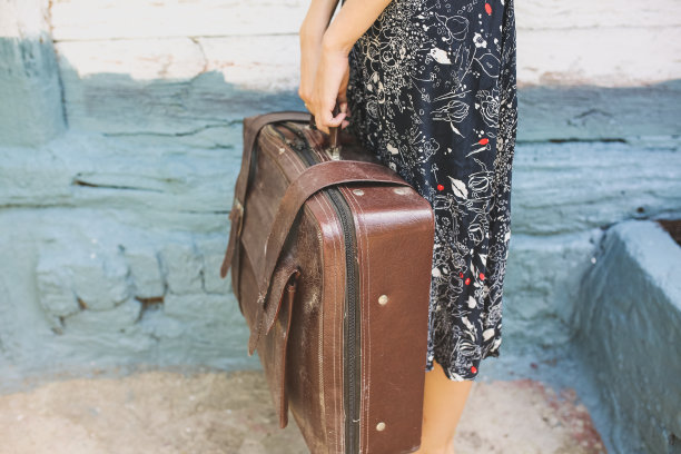 一个带着旧皮箱的时髦女孩站在木墙旁边。时尚的旅游照片。村子里穿着黑衣服的女人。复古乡村的概念。图片下载