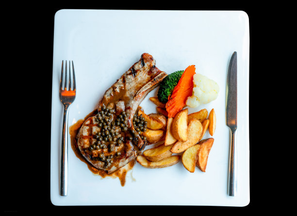 上面的牛排淋上酱汁和胡椒，配上炸土豆、胡萝卜、花椰菜和蒸花椰菜。牛排放在白色盘子里，用不锈钢刀叉。美味的蛋白质食品。图片下载