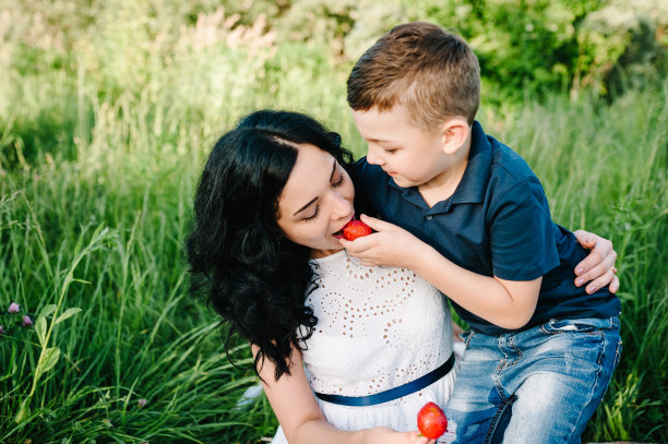 妈妈和儿子吃草莓的照片。快乐的年轻家庭在户外度假。妈妈，小男孩在毯子上野餐，草地，大自然。家庭度假的概念。图片下载