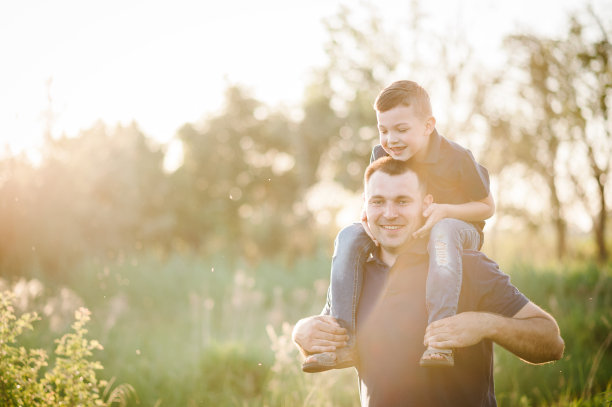 日落时分，小男孩坐在爸爸的肩膀上在公园里玩耍。友好家庭的概念。父亲节。爸爸和儿子在暑假里的照片。放置文字、广告。图片下载