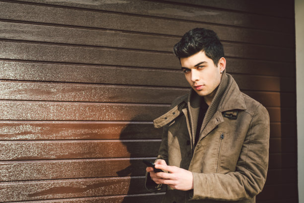 肖像年轻英俊的土耳其男性模特地中海种族深色皮革大衣使用手机的技术商人在街道上在阳光明媚的天气对木墙的背景图片下载